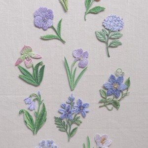 Puede incluir: Una colección de diez parches bordados florales individuales con varios diseños botánicos en tonos morado, lavanda, azul, amarillo y verde. Cada parche muestra un trabajo de hilo detallado que representa diferentes especies de flores.
