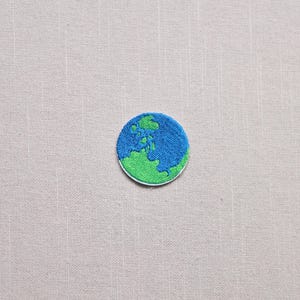 May include: Embroidered patch featuring a circular design of the Earth. The patch has a blue ocean and green landmass, with a white border. The patch is likely for clothing or accessories.