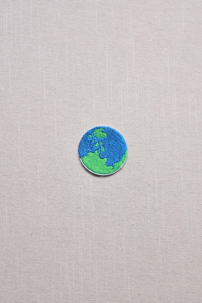 May include: Embroidered patch featuring a circular design of the Earth. The patch has a blue ocean and green landmass, with a white border. The patch is likely for clothing or accessories.
