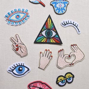 Puede incluir: Una colección de parches bordados con varios diseños. Se incluyen un ojo dentro de un triángulo, una mano haciendo el signo de la paz, una mano formando un corazón, una pluma, una mano de Hamsa, un par de ojos, un par de labios y un ojo.