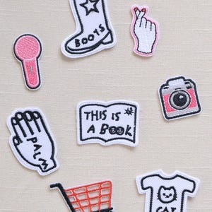 Peut inclure: Ensemble de patchs brodés avec divers motifs. Comprend un microphone, une botte de cowboy avec une étoile, une main en forme de cœur, une main avec "PALM", un livre avec du texte, un appareil photo, un chariot de courses et un t-shirt avec un chat.