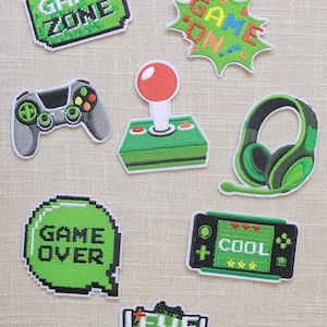 Könnte beinhalten: Eine Sammlung von aufgestickten Patches mit Gaming-Motiven. Designs umfassen einen Joystick, einen Controller, Kopfhörer und Sätze wie "Game Zone", "Game On", "Game Over", "Cool" und "Level Up" in pixeligen Schriftarten. Die Patches sind hauptsächlich grün und grau.