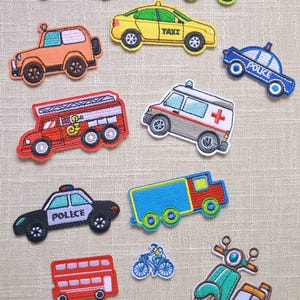May include: A collection of embroidered patches featuring various vehicles including a yellow school bus, a green recycling truck, an orange SUV, a yellow taxi, a blue police car, a red fire truck, an ambulance, a blue truck, a red bus, a bicycle, a scooter, and a yellow submarine.