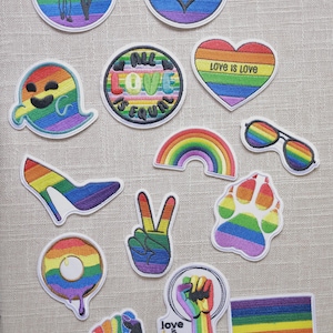 Peut inclure: Divers patchs brodés colorés avec des motifs arc-en-ciel. Les patchs incluent un cœur, des lunettes de soleil, un talon haut, un signe de paix, une empreinte de patte et un beignet. Certains patchs ont le texte "Love is Love" et "All Love is Equal."