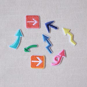 Może przedstawiać: Kolekcja haftowanych naszywek ze strzałkami w różnych kolorach, w tym turkusowym, zielonym, niebieskim, żółtym, różowym, pomarańczowym i czerwonym. Strzałki wskazują w różnych kierunkach, niektóre w kwadratowych ramkach. Naszywki są na neutralnym tle materiału.