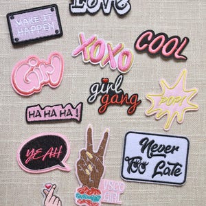 Puede incluir: Una colección de parches bordados con varios diseños y texto. Los parches incluyen frases como "LOVE", "COOL" y "YEAH!", junto con otros gráficos y texto como "XOXO" y "Girl Gang". Los parches son de color rosa, negro y blanco.