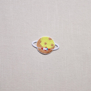 May include: Embroidered patch featuring a planet with a yellow surface and brown ring. A small rocket is embroidered on the planet. The patch is white-bordered and designed for application on clothing or accessories.