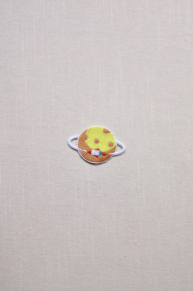 May include: Embroidered patch featuring a planet with a yellow surface and brown ring. A small rocket is embroidered on the planet. The patch is white-bordered and designed for application on clothing or accessories.
