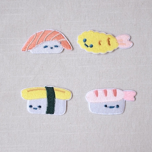 以下が含まれることがあります： 4つの刺繍された寿司パッチ。パッチには、サーモン握り、黄色の天ぷらエビ、緑の海苔ストリップが付いた玉子握り、ピンクのエビ寿司が含まれます。各パッチには笑顔があります。