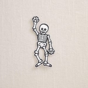 May include: A white and black embroidered skeleton patch. The skeleton is waving with one arm raised. The patch is a fun, spooky accessory for Halloween or any time of year.