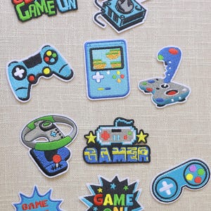 Può includere: Una collezione di toppe ricamate a tema gaming. I disegni includono controller di gioco, un joystick e il testo che dice "Get Your Game On", "Gamer", "Game Over" e "Game On!" in vari colori.