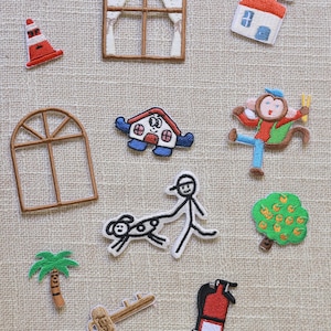 Puede incluir: Una colección de parches bordados que incluye un cono de tráfico, una ventana con cortinas, una casa, un mono, una ventana arqueada, una casa de dibujos animados, un monigote paseando a un perro, un árbol frutal, una palmera, una llave y un extintor.