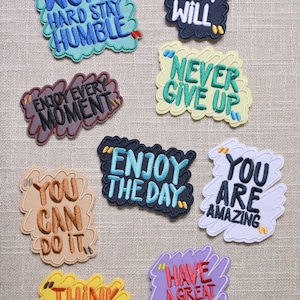May include: A collection of embroidered patches with motivational phrases. The patches feature phrases like "Work Hard Stay Humble," "I Can and I Will," and "Enjoy the Day." Each patch has a unique color scheme and font style.
