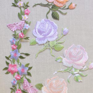 Könnte beinhalten: Gestickte Blumenaufnäher mit Rosen und anderen Blumen in Rosa-, Pfirsich- und Lavendeltönen. Die Aufnäher enthalten grüne Blätter und Stiele sowie einen Schmetterling. Die Aufnäher befinden sich auf beigem Hintergrund.