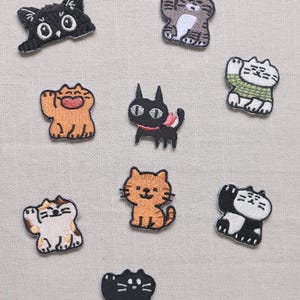 May include: A collection of embroidered cat patches in various colors and poses. The patches include black, orange, gray, and white cats, some waving or sitting. The patches are on a light beige fabric background.