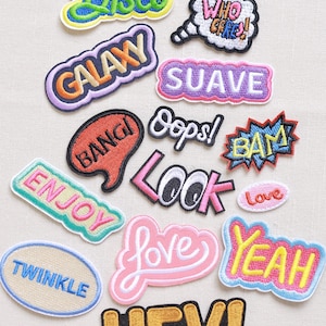 May include: A collection of colourful embroidered iron-on patches with various words and phrases, including "Babe", "Disco", "Who Cares", "Galaxy", "Suave", "Bang", "Oops", "Bam", "Enjoy", "Look", "Love", "Yeah", "Twinkle", "Hey!" and "Love".