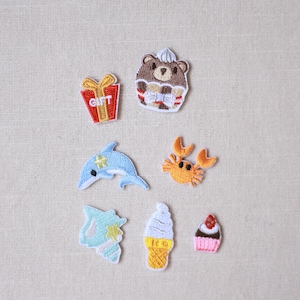 Puede incluir: Colección de parches bordados con varios diseños. Incluye una caja de regalo con el texto "GIFT", un oso con sombrero, un delfín azul, un cangrejo naranja, una concha, un helado y una magdalena. Todos los parches tienen apariencia cosida.