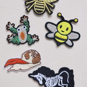 May include: A collection of embroidered patches featuring various designs. Includes a gold bee, a green and white frog, a yellow and black bee, a pug with a red cape, and a black unicorn skeleton. These patches are suitable for clothing or accessories.