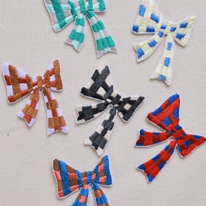 May include: Seven embroidered bow appliques in a variety of colours. The bows have a checkered pattern in shades of green, blue, red, brown, and black. Each bow has a white border and is set against a neutral background.