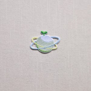 May include: Embroidered patch featuring a planet design with a light blue and yellow orbit. The planet is light green and blue with a small green sprout on top. The patch is on a beige fabric background.
