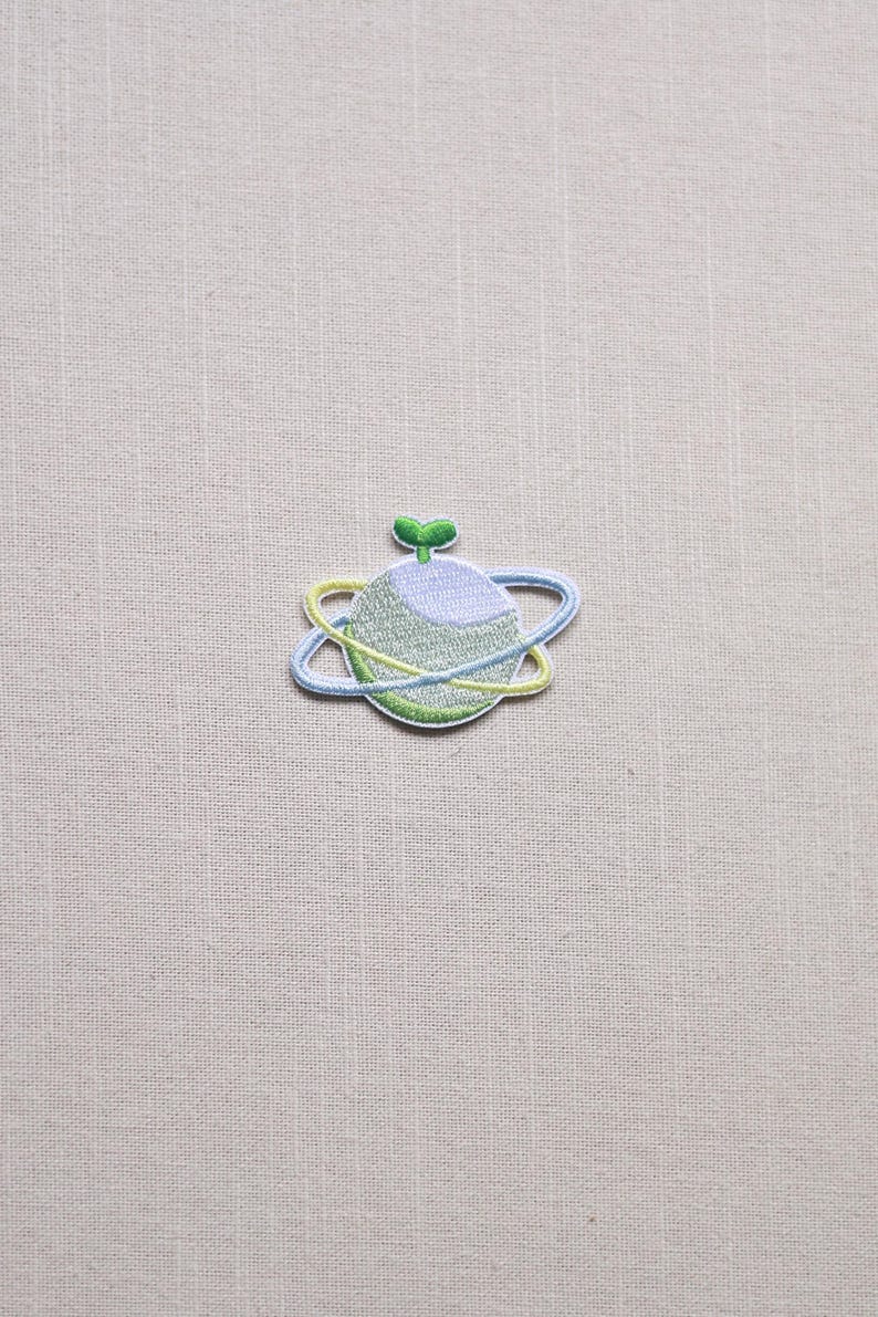 May include: Embroidered patch featuring a planet design with a light blue and yellow orbit. The planet is light green and blue with a small green sprout on top. The patch is on a beige fabric background.