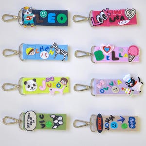May include: Eight rectangular fabric keychains with silver-tone metal lobster clasps. Each strap features colourful embroidered patches, letters, and icons including animals, food, smiley faces, and names like LEO, LISA, CHEN, BELLA, JUNO, OLIVIA, VIC, and JOHN.