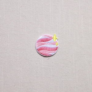 May include: Embroidered patch featuring a pink and white planet design with yellow star accents. The patch is circular and has a detailed, stitched appearance, suitable for clothing or accessories.
