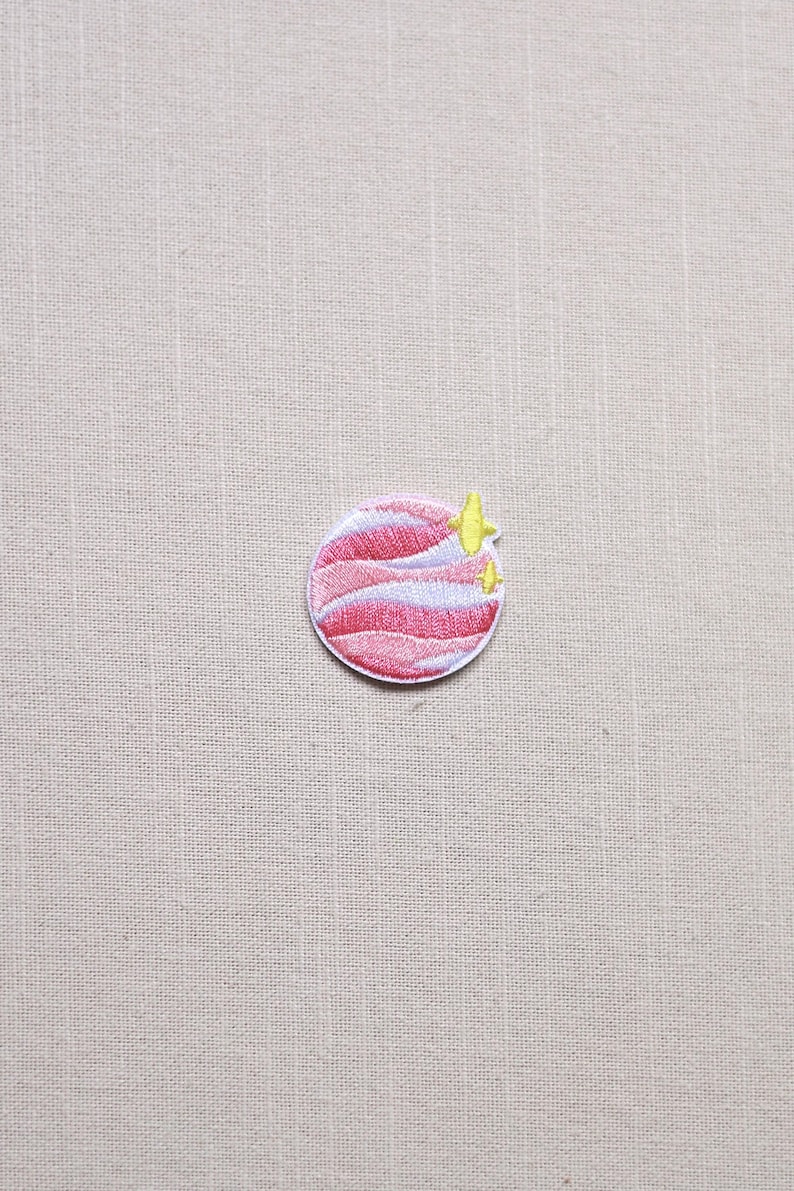 May include: Embroidered patch featuring a pink and white planet design with yellow star accents. The patch is circular and has a detailed, stitched appearance, suitable for clothing or accessories.