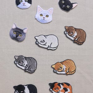 May include: A selection of embroidered cat patches in different colours and poses. The patches feature cat faces and sleeping cat designs in black, white, grey, and orange. These patches are suitable for adding to clothing or accessories.