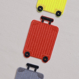 Può includere: Tre toppe ricamate a forma di bagaglio in giallo, rosso e grigio. Ogni toppa presenta un design a valigia con ruote e maniglia nere. Le toppe sono disposte diagonalmente su uno sfondo grigio chiaro.