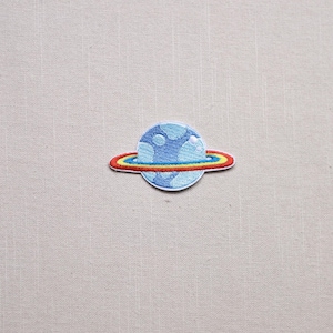 May include: Embroidered patch featuring a blue planet with white cloud-like details and a colorful ring. The ring has red, yellow, and blue bands. The patch is on a light beige fabric background.