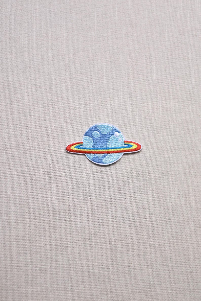 May include: Embroidered patch featuring a blue planet with white cloud-like details and a colorful ring. The ring has red, yellow, and blue bands. The patch is on a light beige fabric background.