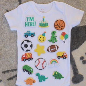 May include: A white short-sleeved baby bodysuit featuring various colorful embroidered patches including a green 'I'M HERE!' text, a birthday cake, basketball, truck, smiley face, dinosaur, balloons, soccer ball, star, football, dump truck, baseball, rainbow, and garbage truck.