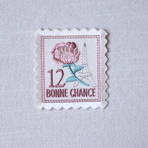 May include: Embroidered patch featuring a pink rose, the Eiffel Tower, and the number 12. The text "BONNE CHANCE" is below the image. The patch has a white scalloped border and is set against a white fabric background.