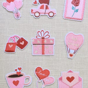 Puede incluir: Una colección de parches bordados en tonos rosa y rojo, con temas de San Valentín. Los diseños incluyen globos en forma de corazón, un coche con un corazón, una rosa, cajas de regalo, un globo aerostático, una taza de café, globos de amor y un sobre.