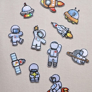 May include: A selection of embroidered space-themed patches. The designs include astronauts, rockets, a satellite, and a UFO. The patches are in various colours, such as blue, orange, and white, and are likely for decorating clothing or accessories.