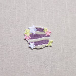 May include: Embroidered patch featuring a purple and white striped design, resembling a layered cake. The patch is adorned with small, colorful flower accents in pink, yellow, and white. The patch is on a neutral fabric background.