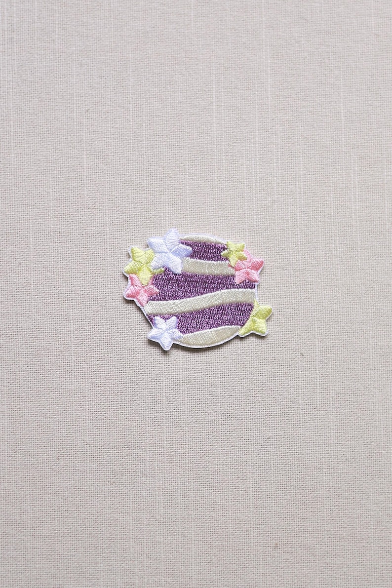 May include: Embroidered patch featuring a purple and white striped design, resembling a layered cake. The patch is adorned with small, colorful flower accents in pink, yellow, and white. The patch is on a neutral fabric background.