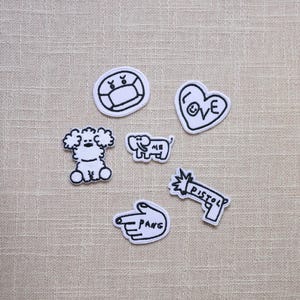 May include: A collection of six white and black embroidered patches. Designs include a face wearing a mask, a heart with "LOVE", a poodle, an elephant with "ME", a hand pointing with "PANG", and a pistol with "PISTOL".