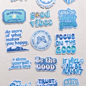 May include: A collection of fifteen embroidered patches in various shades of blue and white. Each patch features unique motivational text and graphic elements, including flowers, butterflies, hearts, and suns, with phrases like Stay Happy, Good Vibes, and Trust Your Visions.