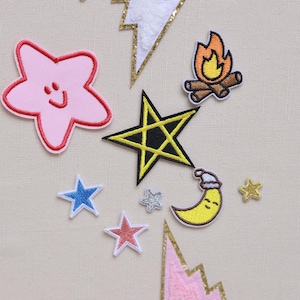 Könnte beinhalten: Eine Sammlung von gestickten Aufnähern in verschiedenen Formen und Farben. Enthält Sterne, Blitze, ein Lagerfeuer und einen Mond mit Weihnachtsmütze. Die Aufnäher haben goldene Glitzerakzente auf neutralem Hintergrund.
