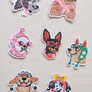 May include: Seven embroidered iron-on patches featuring dogs in various accessories. One patch shows a grey dog in a pink hat with the word Nice. Others include a brown dog in a pink headscarf, a white dog in a pink bonnet, a black dog with red glasses, a tan dog in a green hood, a golden dog with goggles, and a dalmatian with a yellow bow.