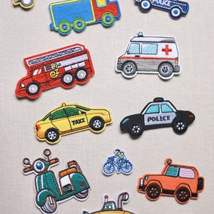 May include: A collection of embroidered patches featuring various vehicles. Includes a yellow school bus, a blue and red truck, a police car, a fire truck, an ambulance, a taxi, a black police car, a scooter, a bicycle, an orange jeep, a submarine, and a red double-decker bus.