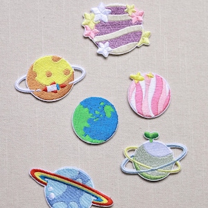 May include: A collection of embroidered planet patches. The patches feature various planets with colorful designs, including a planet with a rocket, a striped planet, and a planet with a rainbow ring. The patches are on a neutral background.