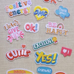 Op de afbeelding: Een verzameling geborduurde patches met positieve berichten en zinnen. Ontwerpen zijn onder meer "You're Doing Well", "Positive Energy", "I Will Be OK", "Thank You", "Never Give Up", "OK!", "Be Happy", "Cutie", "Excellent", "Yes!", "Friends!", "Beach Bum" en "Oooops!."