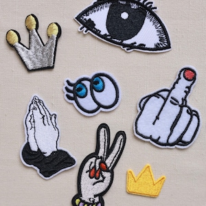 May include: A selection of embroidered patches with diverse designs. These include a silver crown with gold detailing, an eye, praying hands, two cartoon eyes, a hand giving the middle finger, a peace sign hand, and a small yellow crown.