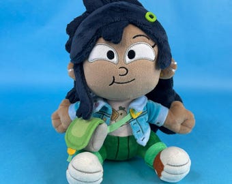 May include: Plush doll of a cartoon character with dark hair, wearing a blue jacket, green pants, and white shoes. The doll has a small green bag and earrings. The background is a solid light blue.