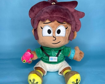 May include: A plush doll of a cartoon character with brown skin, wearing a green hoodie, white shorts, and brown shoes with yellow accents. The doll has brown hair, large eyes, and is holding a pink toy. A blue background.
