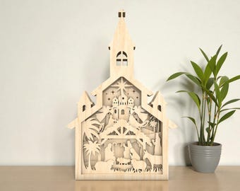 May include: A wooden nativity scene, laser-cut with intricate details. The design features a church-like structure with a steeple, housing a layered depiction of the nativity, including figures, animals, and stars. The natural wood color provides a warm, neutral tone.
