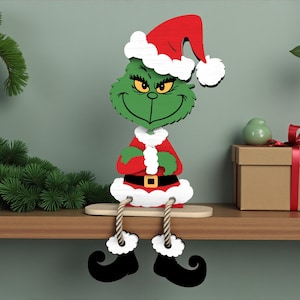 May include: A wooden Christmas decoration depicting a green character in a red Santa hat and coat, with black boots and a sly smile. The figure is designed to sit on a shelf, with dangling legs. The background includes a shelf, a gift box, and festive greenery.